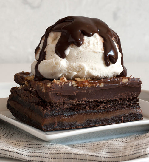 Nut-topped fudge brownie on a white square plate on top of a black and white linen napkin. The fudge brownie is topped with a scoop of vanilla ice cream drizzled in Deep Dark Haute Fudge.